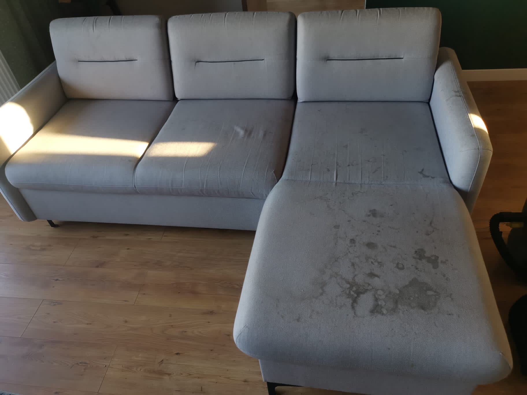 Szara kanapa z widocznymi plamami na siedzisku, wymagająca czyszczenia, na tle drewnianej podłogi.