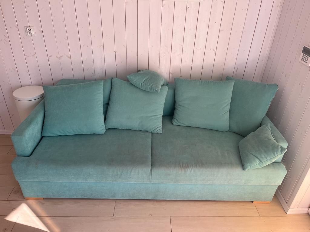 Turkusowa kanapa z poduszkami w pokoju z boazerią, widoczny również biały kosz na śmieci i panel alarmowy.