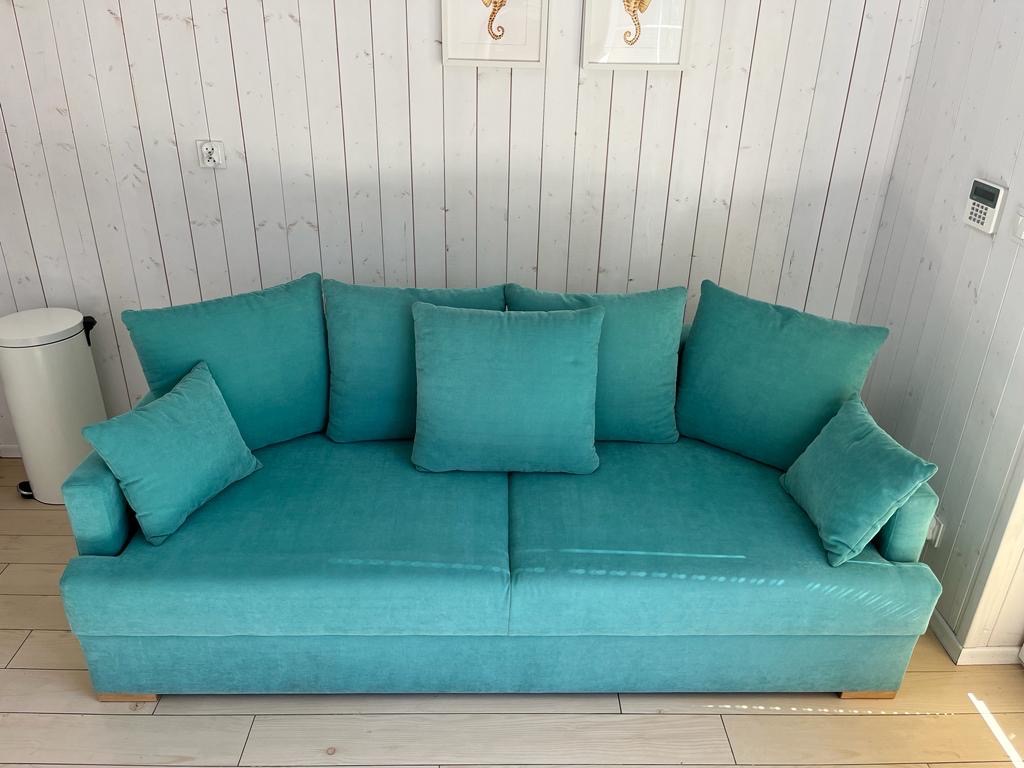 Turkusowa sofa z poduszkami na tle białej, drewnianej ściany z dekoracjami w ramkach przedstawiającymi koniki morskie.