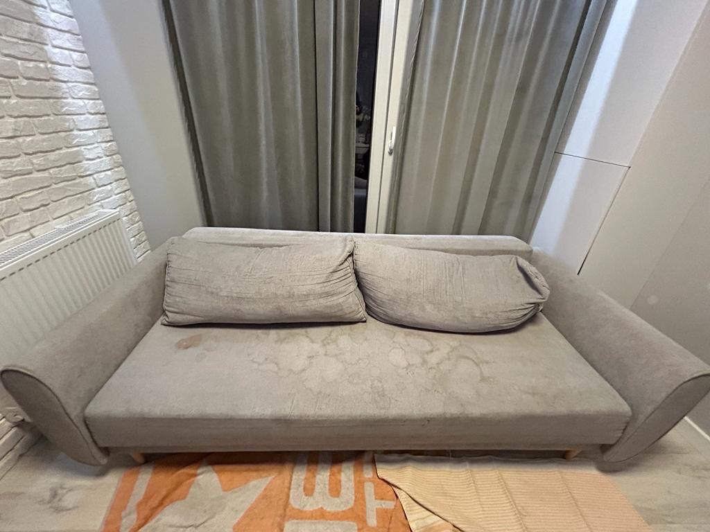 Szara kanapa z plamami na siedzisku i poduszkach, ustawiona pod oknem z zasłonami, widoczna część grzejnika i ściany z cegły.