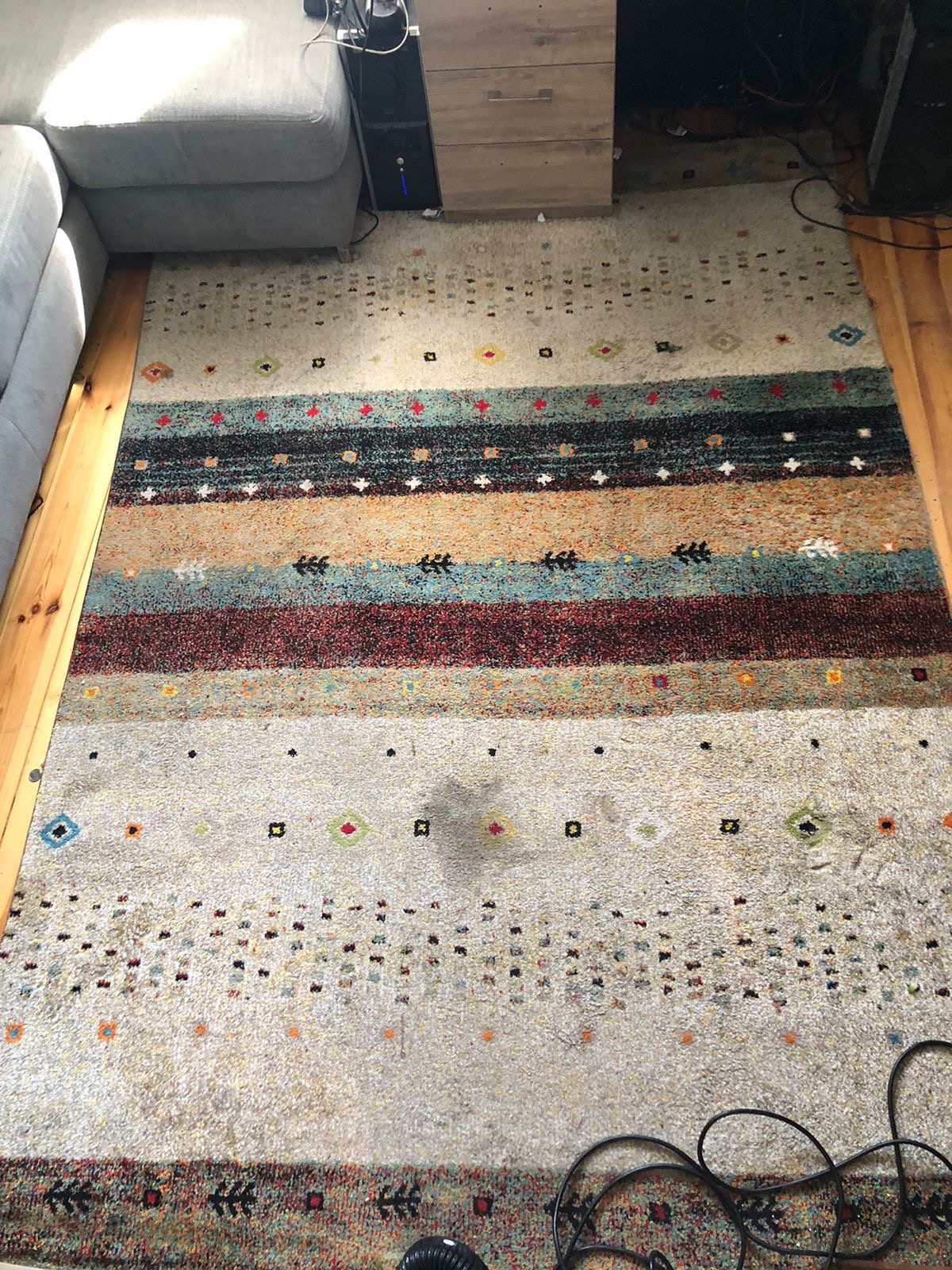 Wielobarwny dywan z geometrycznym wzorem, widoczna plama i wąż od odkurzacza w dolnym rogu.