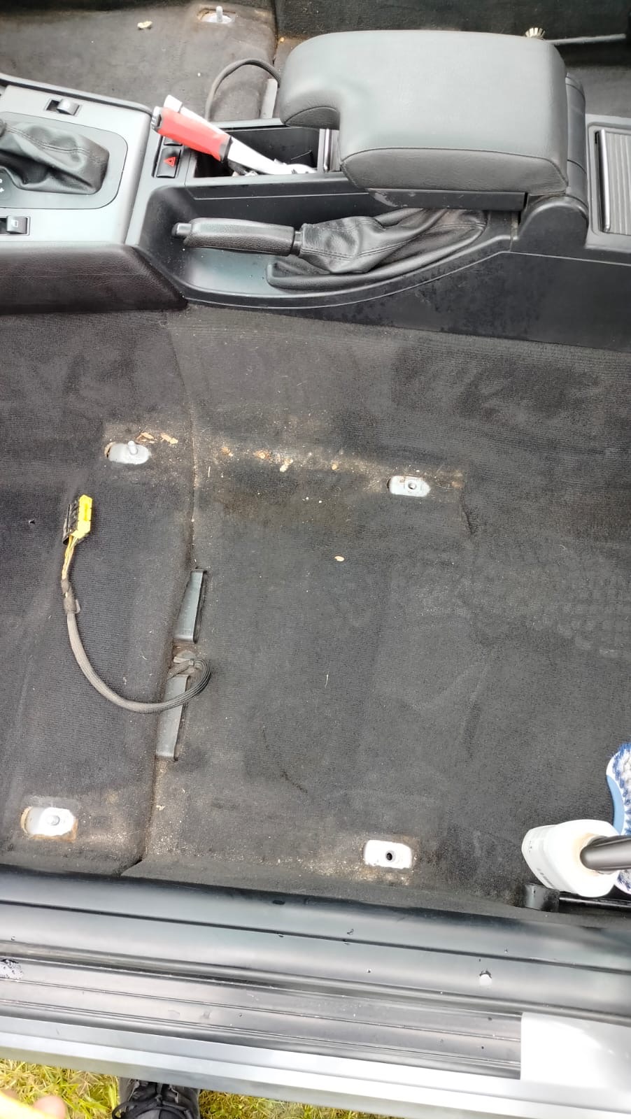 Brudna, czarna wykładzina podłogowa w samochodzie przed czyszczeniem, widoczne mocowania foteli i kabel elektryczny.