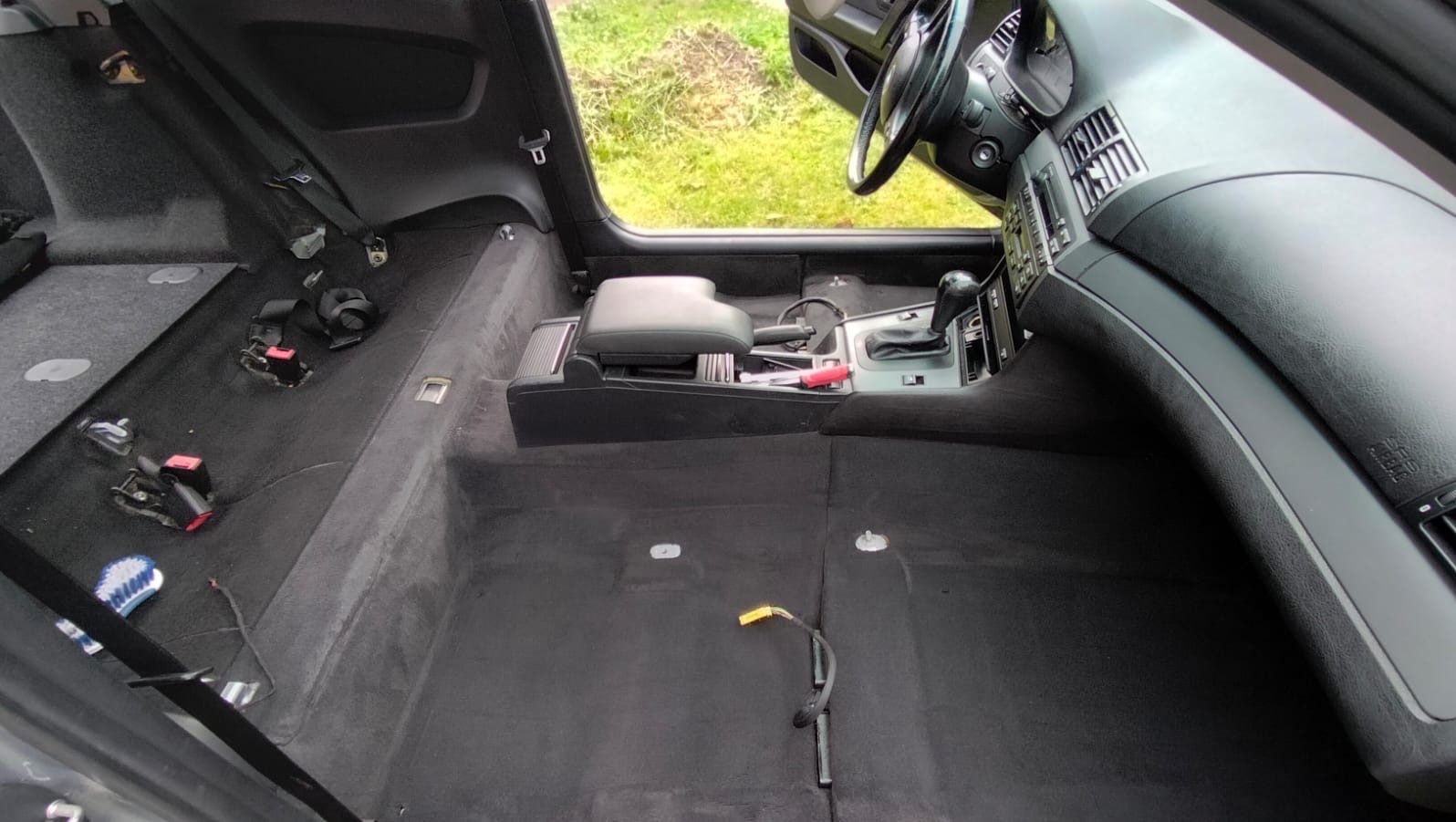 Wnętrze samochodu z wyjętymi siedzeniami, przygotowane do czyszczenia tapicerki podłogowej, widoczne pasy bezpieczeństwa, narzędzia i szczotka.