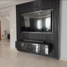 Damian Baran - Nowoczesna ściana telewizyjna z czarnych, pionowych lameli drewnianych, podwieszana szafka RTV z połyskującymi frontami, odbicie okna w ekranie telewizora.