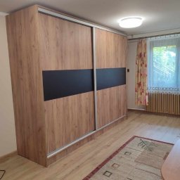 Damian Baran - Duża szafa wnękowa z przesuwnymi drzwiami, okleina w kolorze drewna z czarnym pasem, widoczna w pokoju z dywanem i oknem z zasłonami.