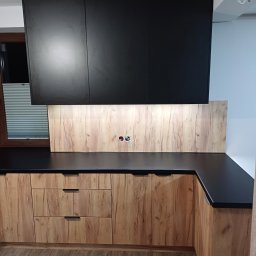 Damian Baran - Nowoczesna kuchnia: czarne szafki wiszące, drewnopodobne fronty dolne, czarny blat, drewniana ściana nad blatem z gniazdkami, widoczne rolety w oknie.