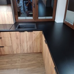 Damian Baran - Kuchenna zabudowa z drewnianymi frontami szafek i czarnym blatem, widoczny zlew i okno z odbiciem osób.