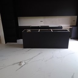 Damian Baran - Częściowo zmontowane czarne szafki kuchenne z blatem roboczym, widoczne gniazdka elektryczne w ścianie, podłoga z białego marmuru, w trakcie montażu.