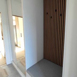 Damian Baran - Wnęka w przedpokoju z siedziskiem i panelem ściennym z drewnianych listew z wieszakami, obok szafa przesuwna z lustrem.