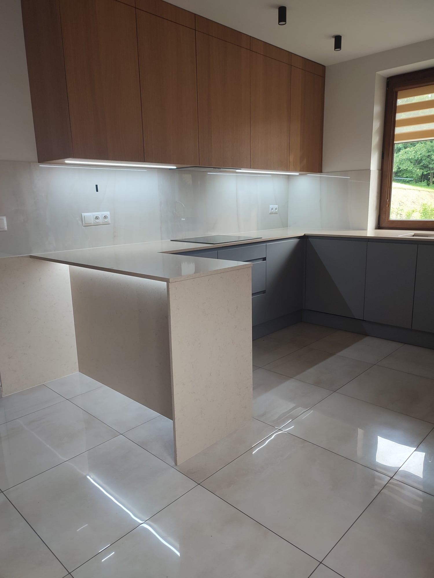 Nowoczesna kuchnia z wyspą, fronty szafek w odcieniach szarości i drewna, podświetlane blaty, płytki podłogowe z połyskiem, okno z widokiem na zieleń.