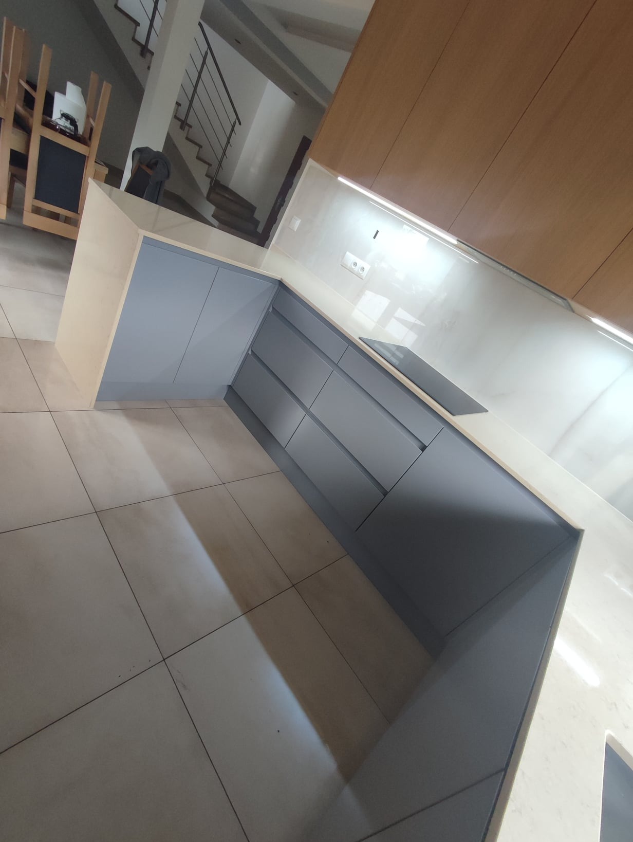 Szare, matowe szafki kuchenne z jasnym blatem, widoczne z niskiego kąta, z elementami płyty indukcyjnej. W tle schody i część jadalni.