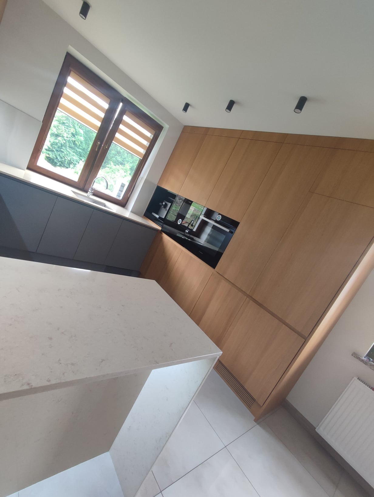 Nowoczesna kuchnia z drewnianymi frontami szafek, szarymi dolnymi szafkami, jasnym blatem wyspy i oknem z żaluzjami poziomymi.