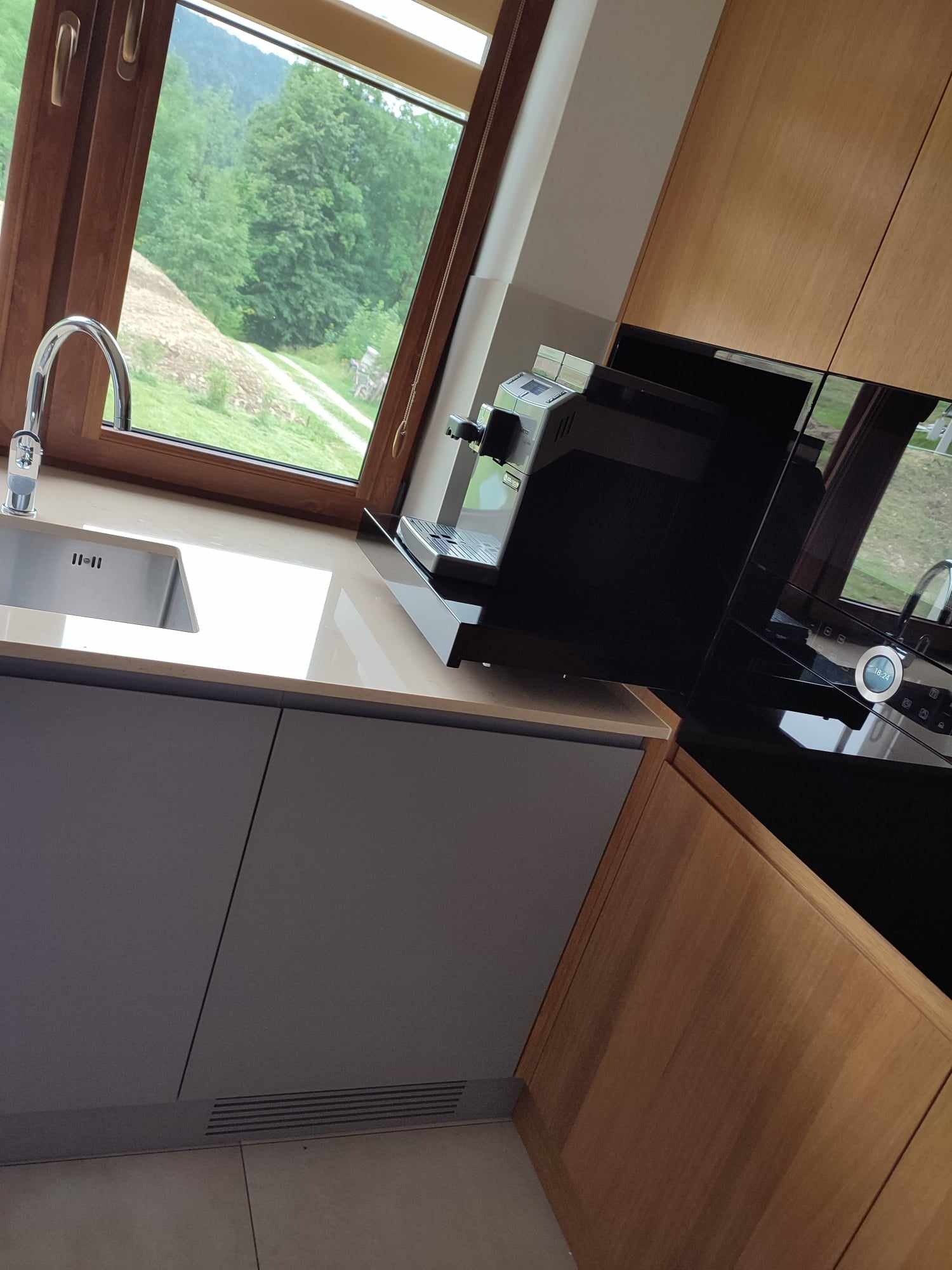 Nowoczesna kuchnia z drewnianymi i szarymi szafkami, widok na zieleń za oknem, ekspres do kawy na blacie.