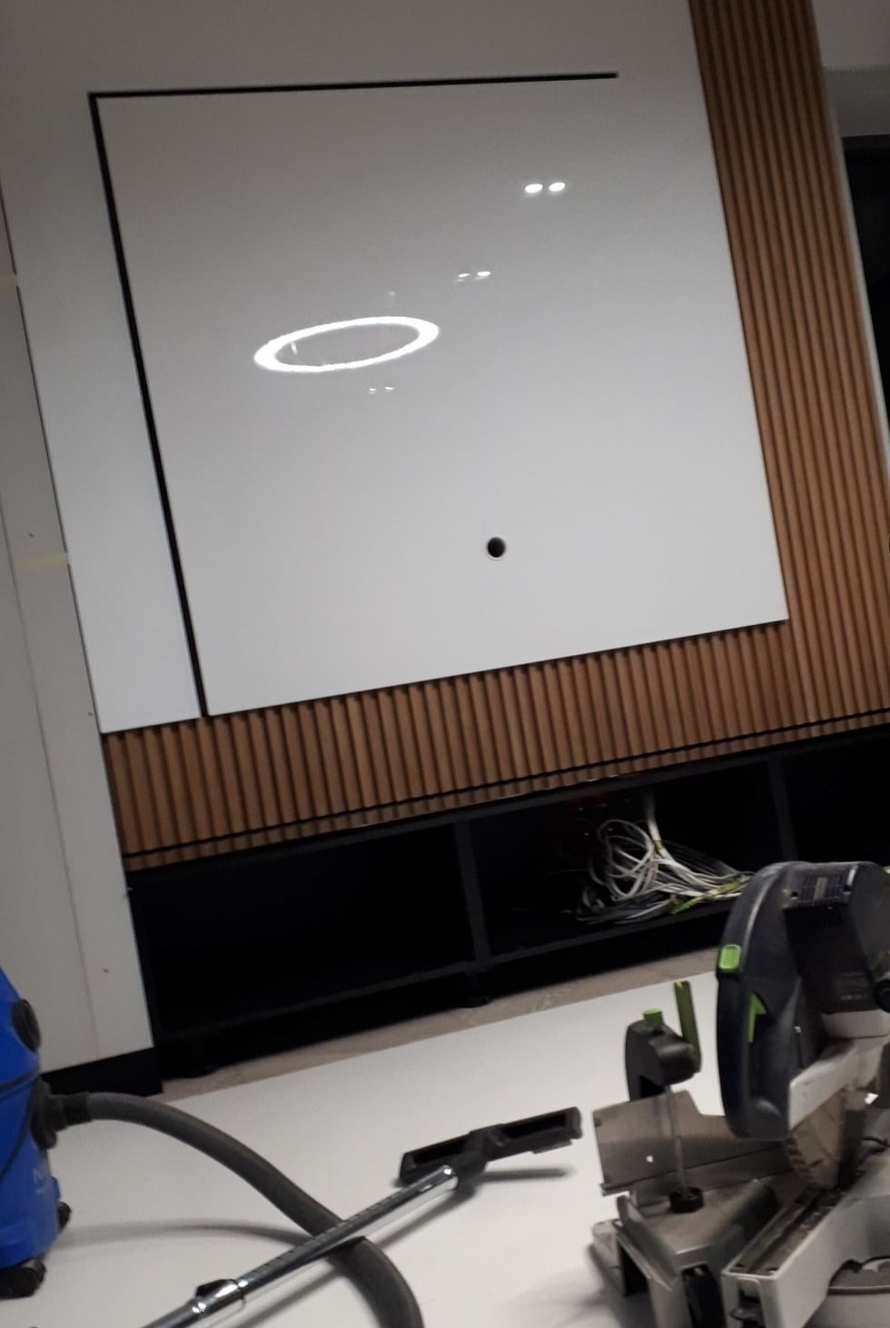 Montaż białej szafy wnękowej z drewnianymi panelami dekoracyjnymi, widoczny odkurzacz i piła ukośnica.