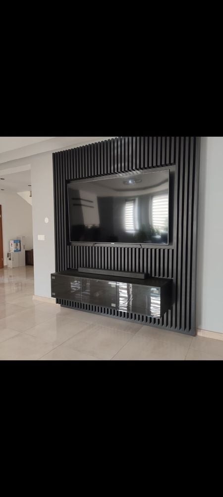 Nowoczesna ściana telewizyjna z czarnych, pionowych lameli drewnianych, podwieszana szafka RTV z połyskującymi frontami, odbicie okna w ekranie telewizora.