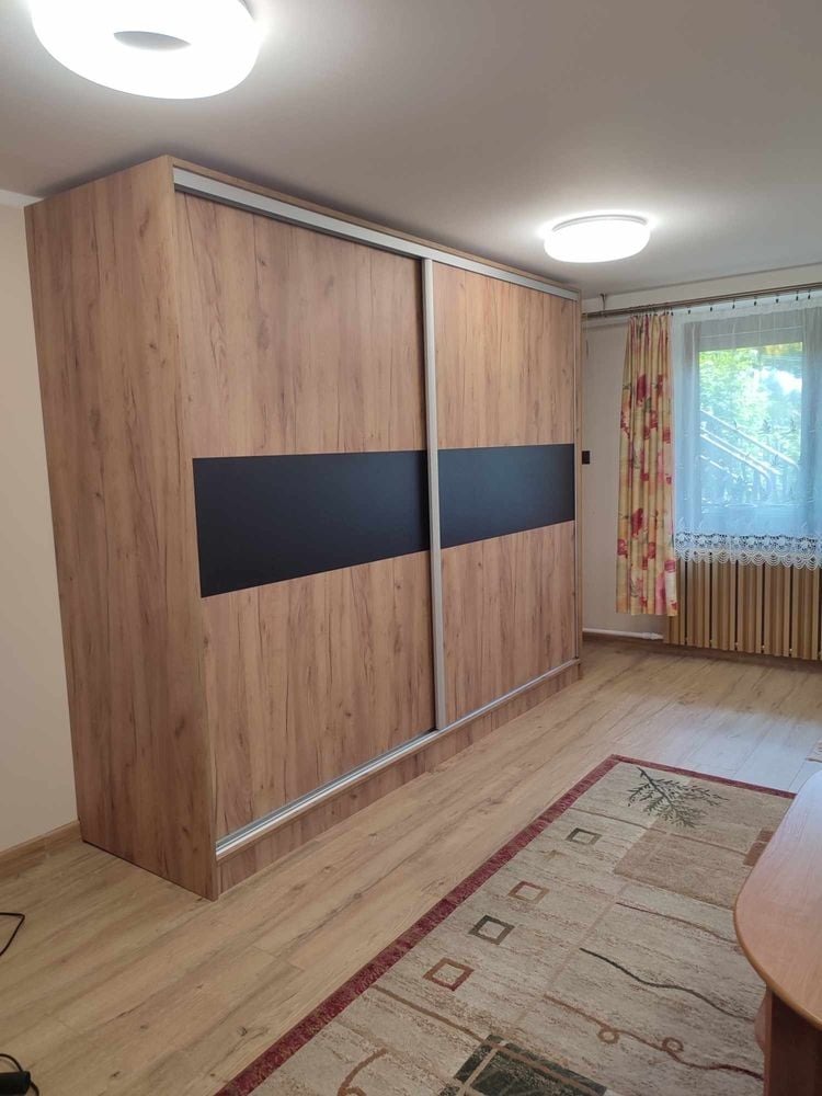 Duża szafa wnękowa z przesuwnymi drzwiami, okleina w kolorze drewna z czarnym pasem, widoczna w pokoju z dywanem i oknem z zasłonami.