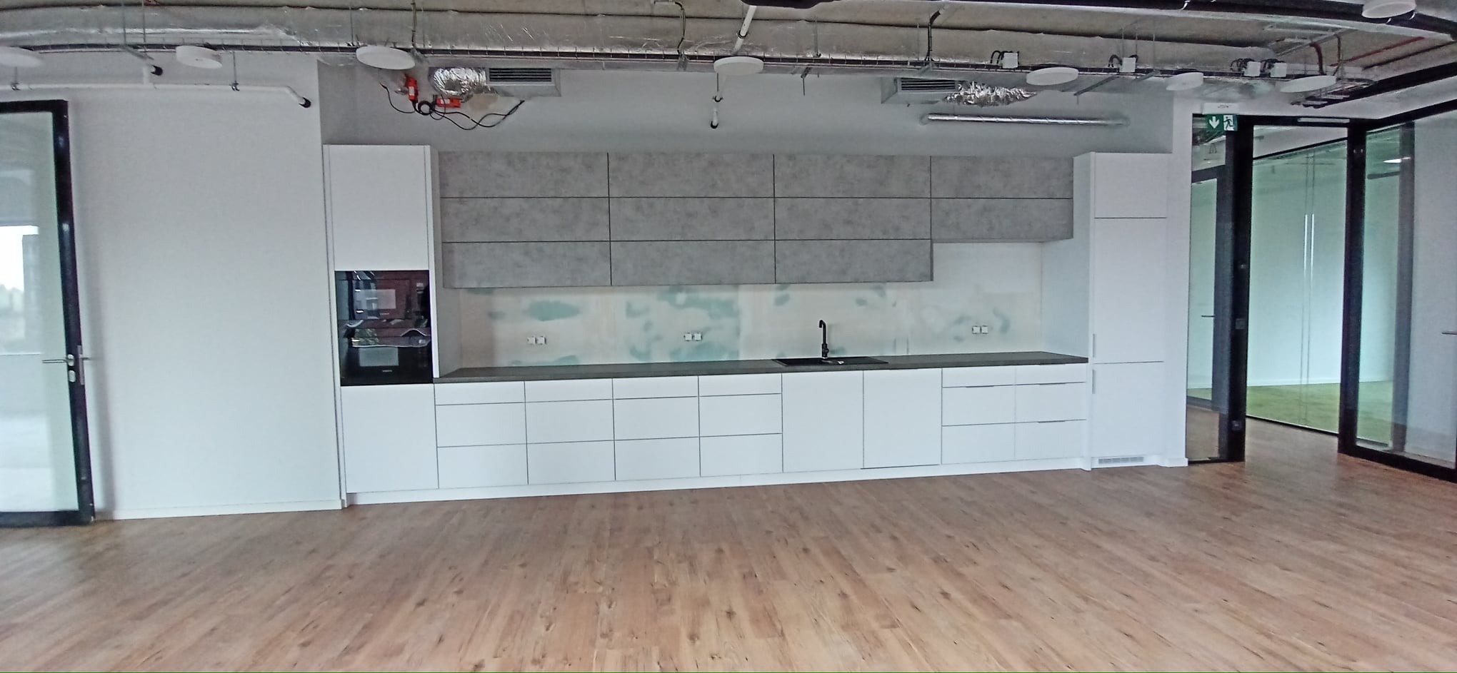 Nowoczesna, minimalistyczna kuchnia z białymi szafkami dolnymi i szarymi górnymi, czarny zlew i armatura, widoczne instalacje pod sufitem w biurowym otoczeniu.