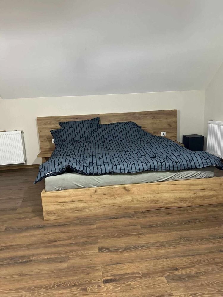 Drewniane łóżko z zagłówkiem i szafkami nocnymi w Nawojowej, przykryte pościelą w geometryczny wzór, na podłodze panele imitujące drewno.