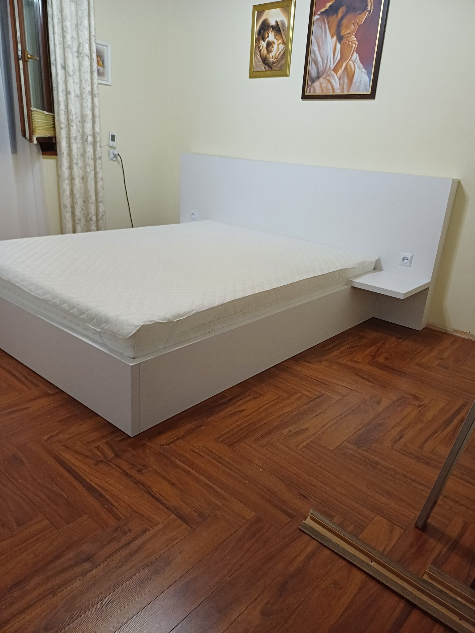 Białe łóżko z materacem na drewnianej podłodze w jodełkę, z widocznymi fragmentami listew przypodłogowych i gniazdkiem elektrycznym wbudowanym w zagłówek.