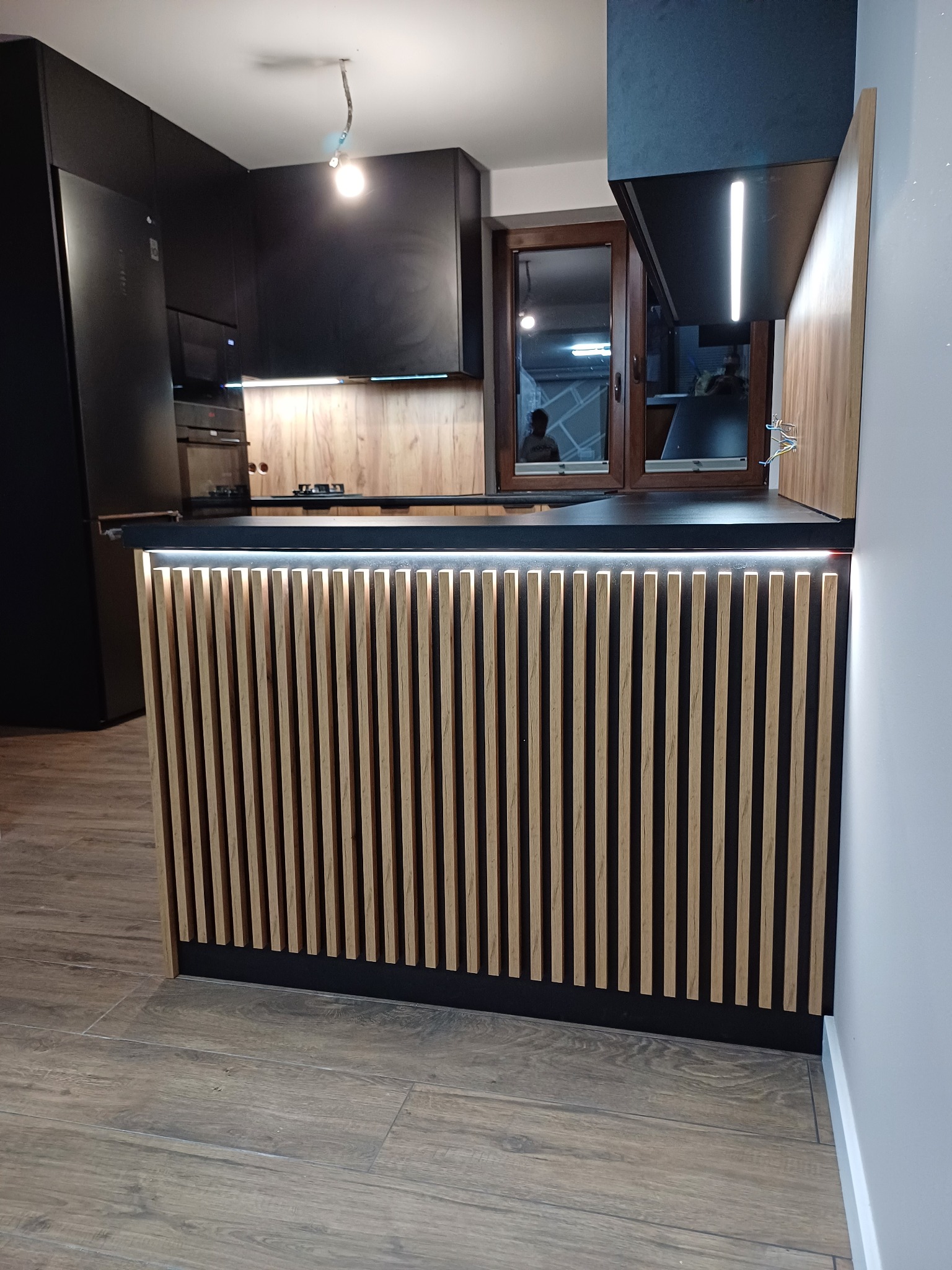 Czarna kuchnia na wymiar z drewnianym wykończeniem wyspy i oświetleniem LED, widok na okno z odbiciem.