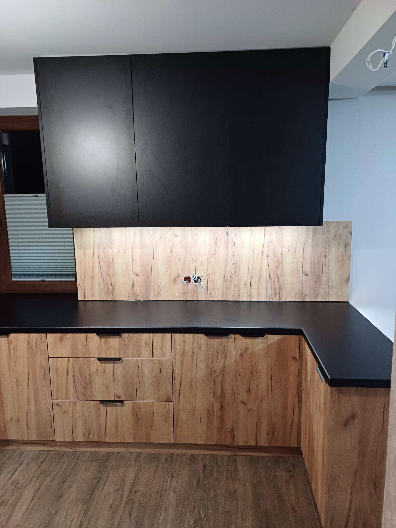 Nowoczesna kuchnia: czarne szafki wiszące, drewnopodobne fronty dolne, czarny blat, drewniana ściana nad blatem z gniazdkami, widoczne rolety w oknie.