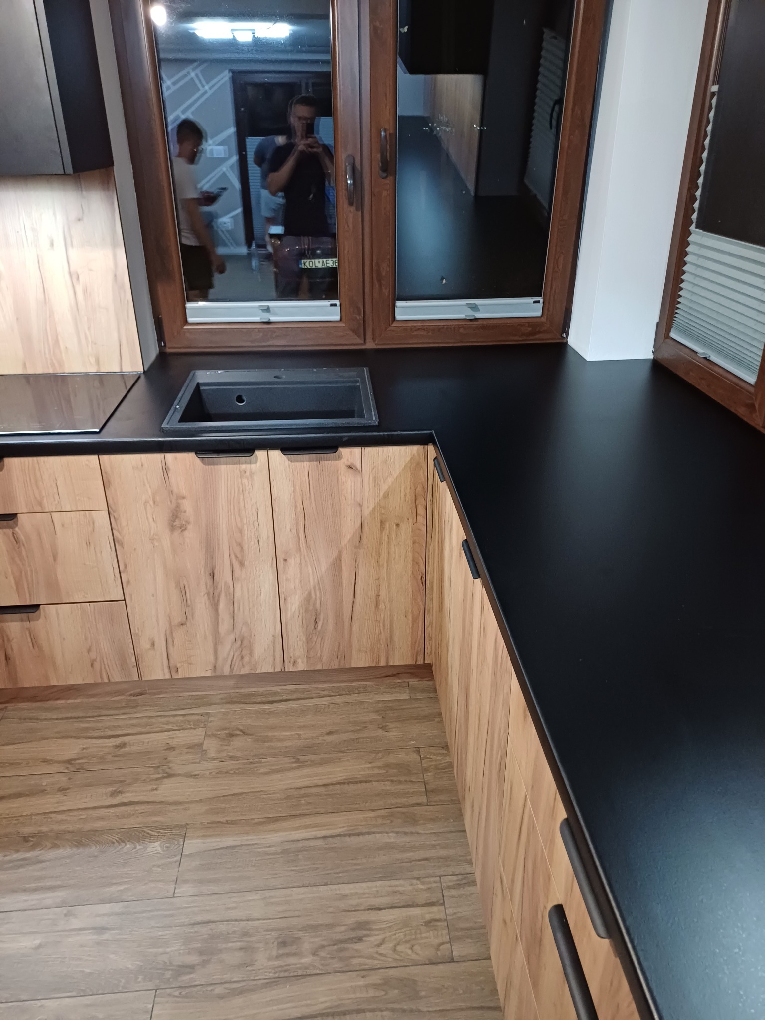 Kuchenna zabudowa z drewnianymi frontami szafek i czarnym blatem, widoczny zlew i okno z odbiciem osób.
