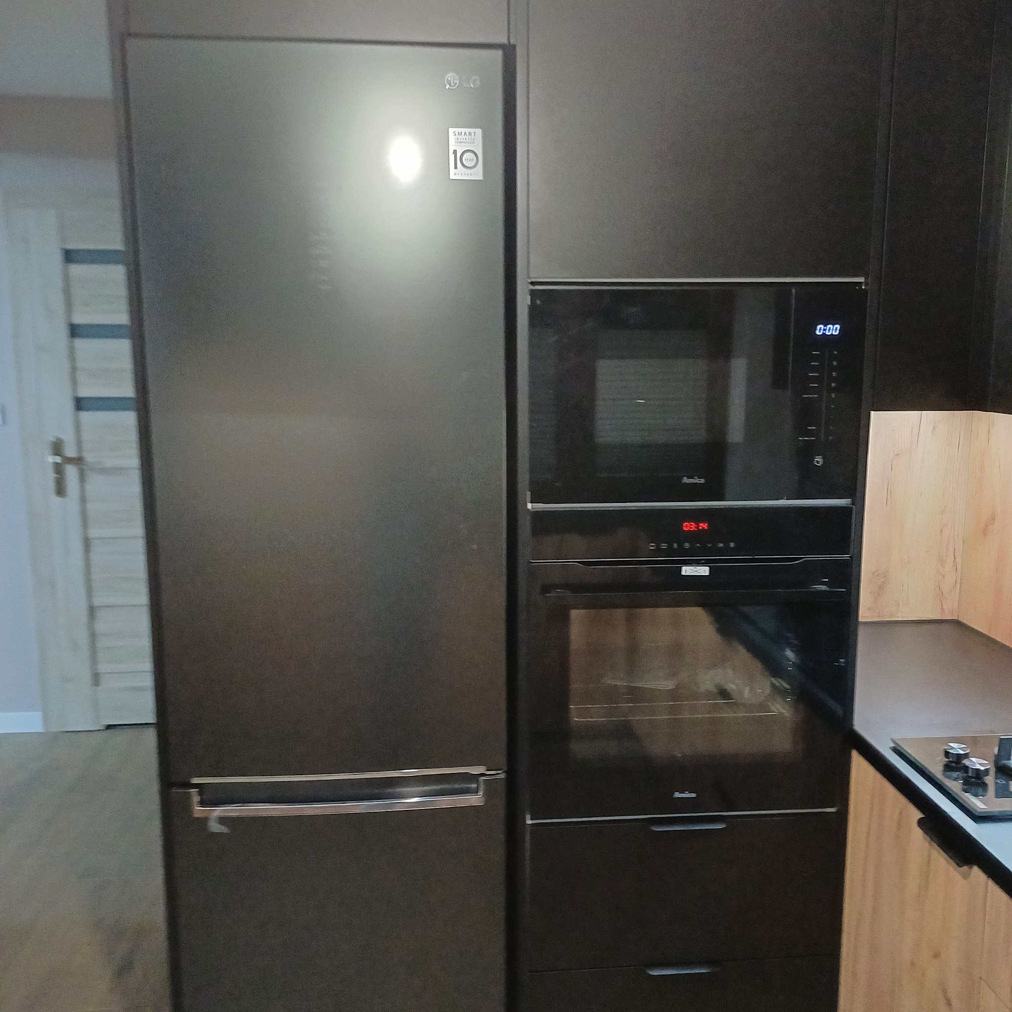 Ciemna zabudowa kuchenna z lodówką LG, piekarnikiem i mikrofalówką Amica, z drewnopodobnym blatem i płytą grzewczą