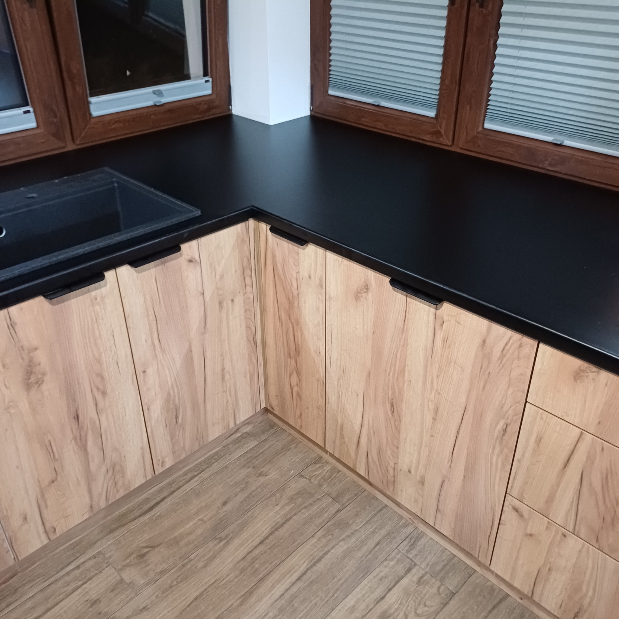 Kąt kuchni z drewnopodobnymi frontami szafek, czarnym blatem i zlewozmywakiem, widok na okno z żaluzjami.