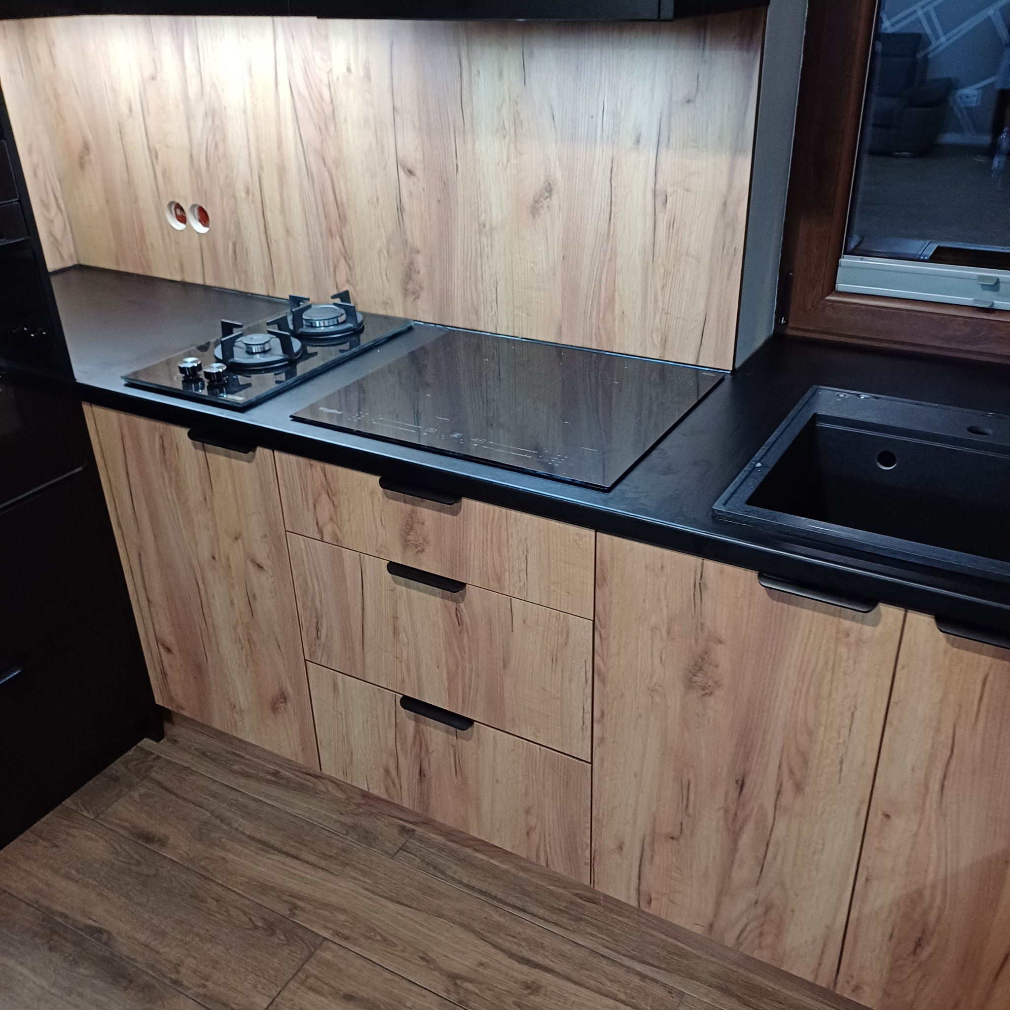 Nowoczesna zabudowa kuchenna z frontami w kolorze drewna, czarnym blatem, płytą indukcyjną i gazową oraz zlewozmywakiem, widoczne gniazdka elektryczne.