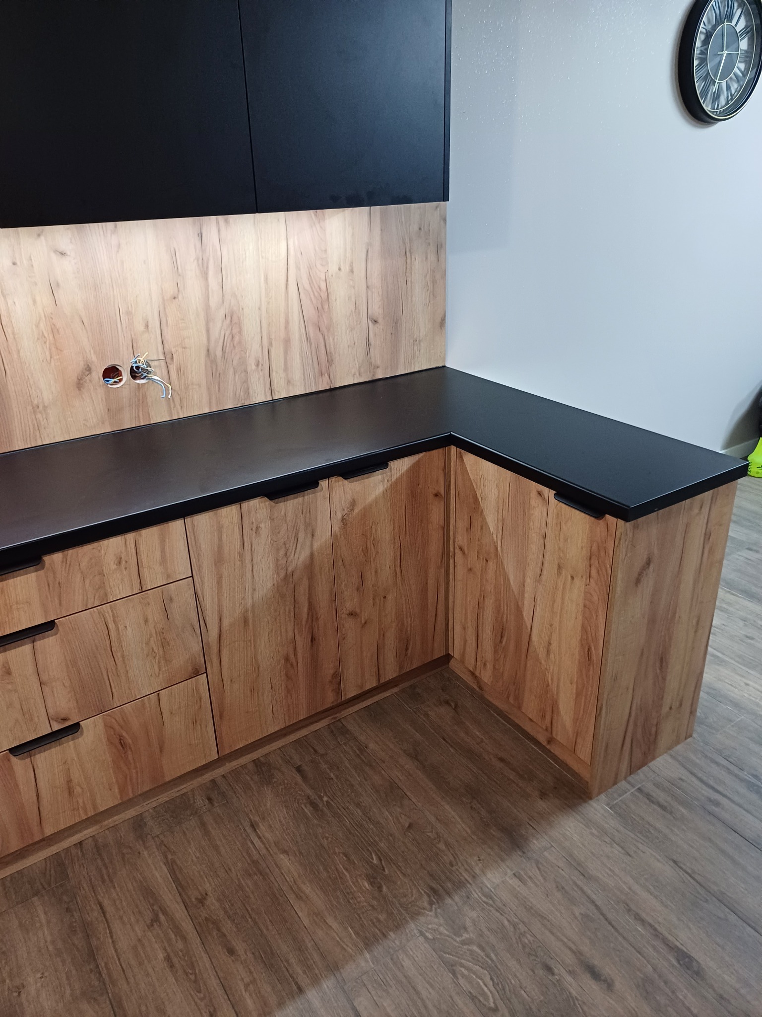 Narożna zabudowa kuchenna z drewnopodobnymi frontami, czarnym blatem i górnymi szafkami, widoczne przygotowane gniazdka elektryczne nad blatem.