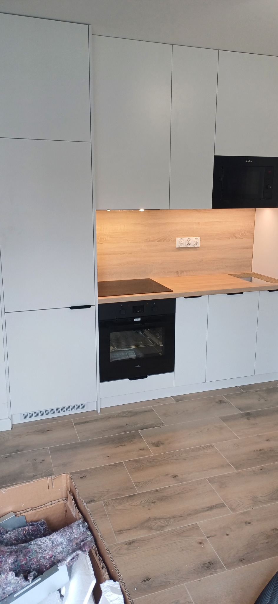 Nowoczesna kuchnia z białymi szafkami, drewnianym blatem i czarnym sprzętem AGD Amica, widoczny fragment kartonu na podłodze.