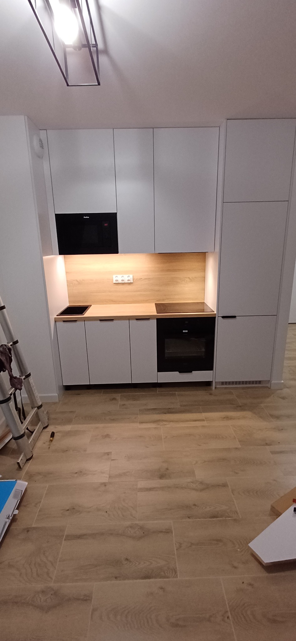 Nowoczesna, biała zabudowa kuchenna z drewnianym blatem i czarnymi sprzętami AGD, widoczna drabina malarska sugeruje trwający montaż.