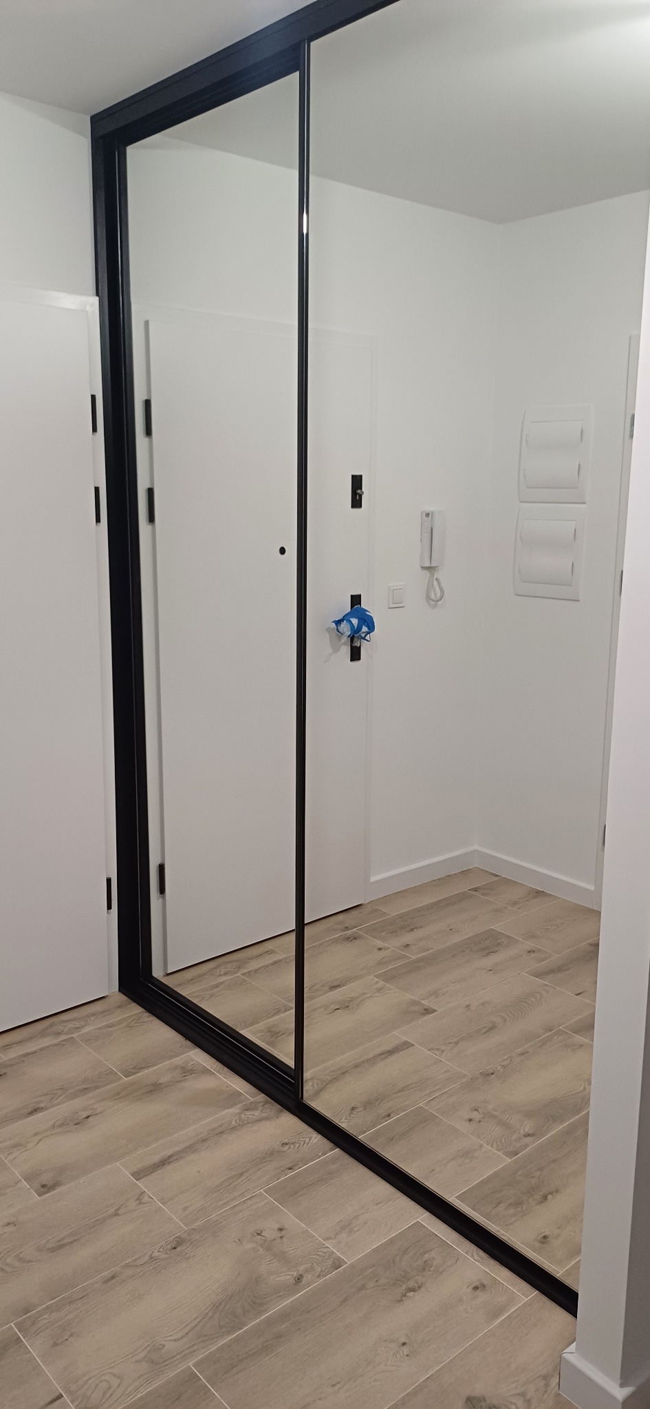 Nowoczesna szafa wnękowa z przesuwnymi drzwiami lustrzanymi i czarną ramą, widok na parkiet z płytek imitujących drewno.