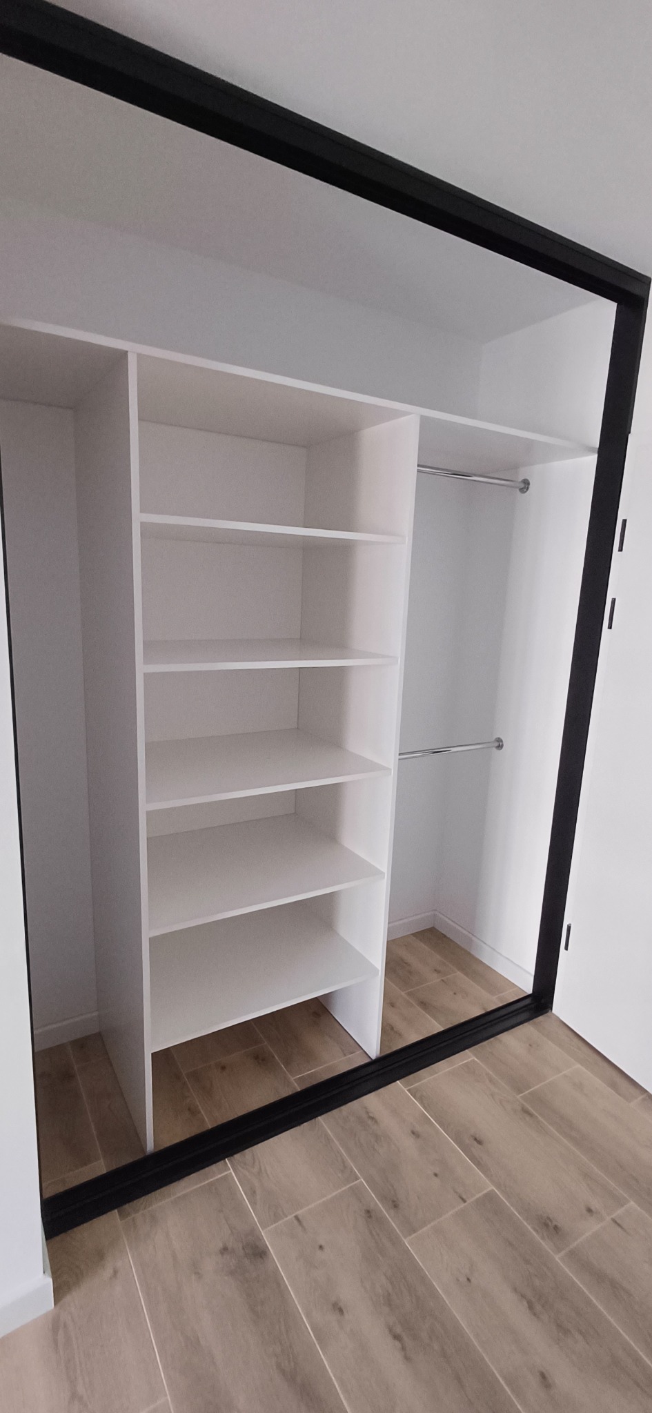 Wnętrze szafy wnękowej z białymi półkami i drążkami, czarna rama drzwi przesuwnych, podłoga wyłożona płytkami imitującymi drewno.