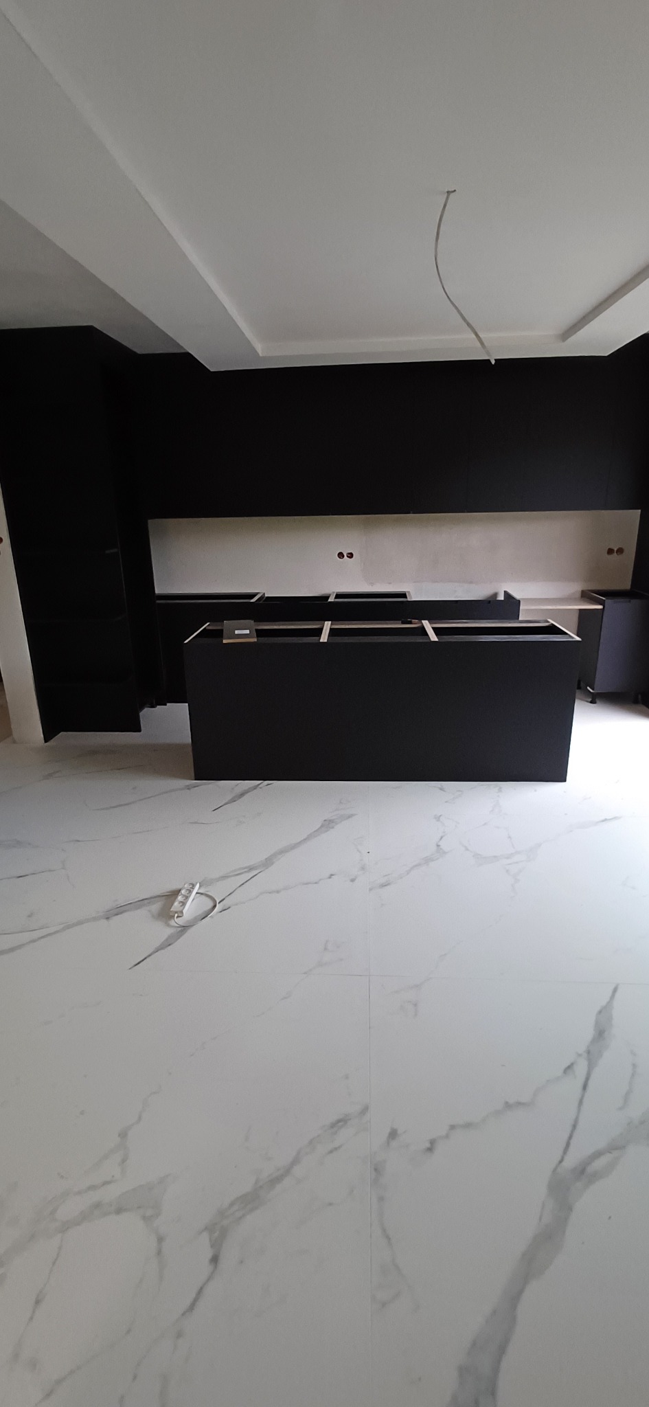 Częściowo zmontowane czarne szafki kuchenne z blatem roboczym, widoczne gniazdka elektryczne w ścianie, podłoga z białego marmuru, w trakcie montażu.