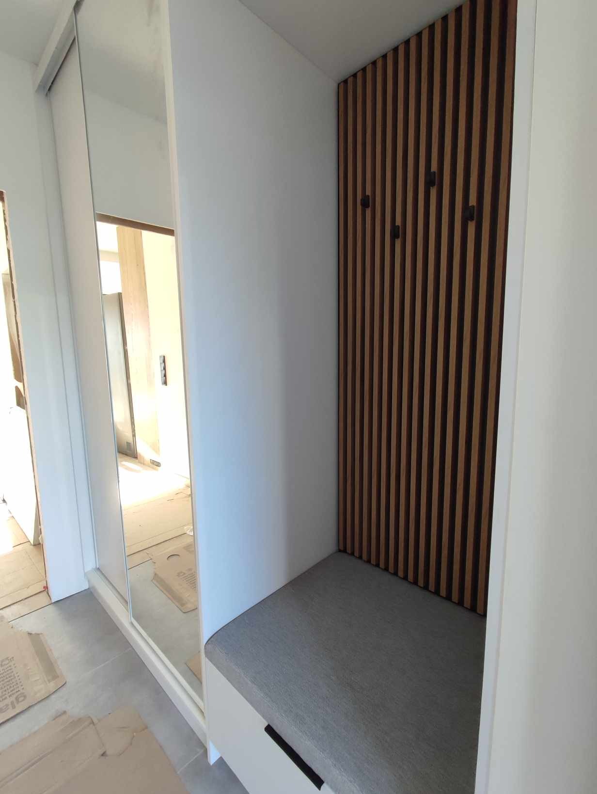 Wnęka w przedpokoju z siedziskiem i panelem ściennym z drewnianych listew z wieszakami, obok szafa przesuwna z lustrem.