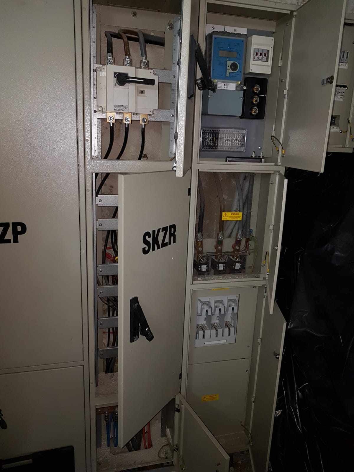 Otwarta skrzynka elektryczna SKZR z widocznymi przewodami, bezpiecznikami i licznikiem energii elektrycznej. Widoczne elementy instalacji elektrycznej.