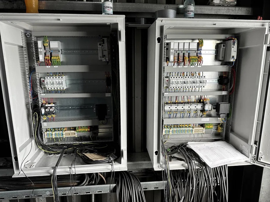 Dwa otwarte panele elektryczne z widocznymi wewnętrznymi komponentami, przewodami i dokumentacją techniczną na jednym z paneli, butelki wody na górze obudów.