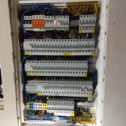 Rozdzielnica elektryczna wnękowa z licznymi bezpiecznikami, zabezpieczeniami różnicowoprądowymi i przewodami, widoczne oznaczenia na urządzeniach.