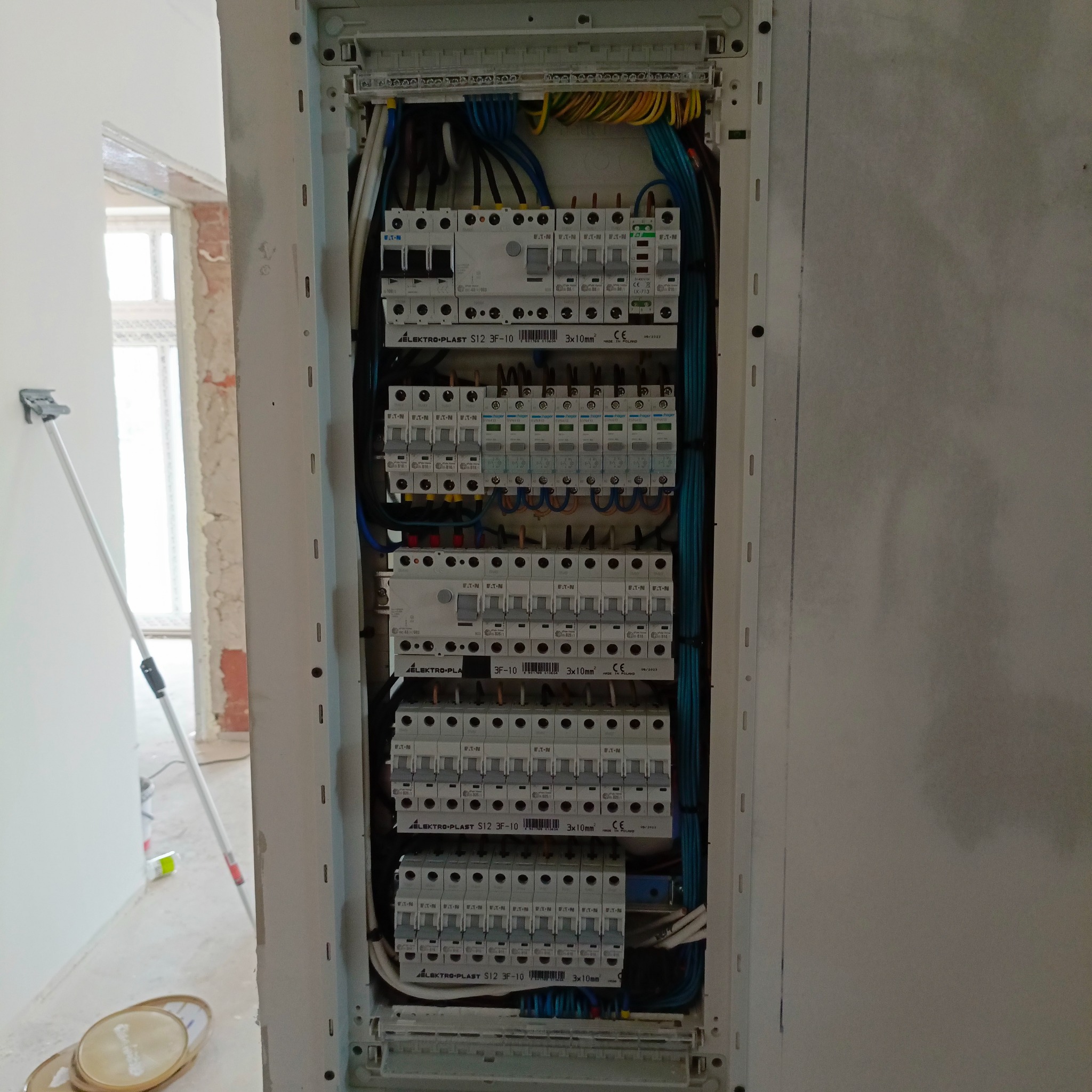 Rozdzielnica elektryczna z zamontowanymi bezpiecznikami Eaton i przewodami Elektro-Plast S12 3F-10, widoczna w trakcie prac wykończeniowych, z gruntowanymi ścianami i narzędziami malarskimi w tle.