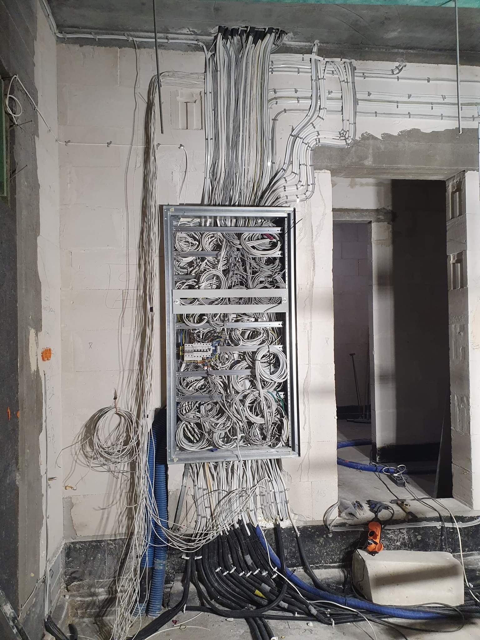 Rozdzielnia elektryczna w trakcie montażu w surowym wnętrzu, widoczne liczne przewody i kable w ścianie z betonu komórkowego, instalacja elektryczna w trakcie wykonywania.