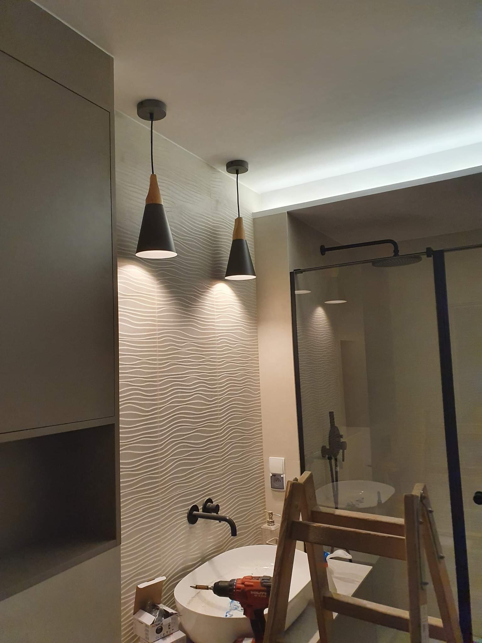Łazienka w trakcie remontu: widoczne dwie lampy wiszące z czarnymi kloszami i drewnianymi elementami, umywalka z czarną armaturą, wiertarka na brzegu umywalki, drabina oparta o kabinę prysznicową...
