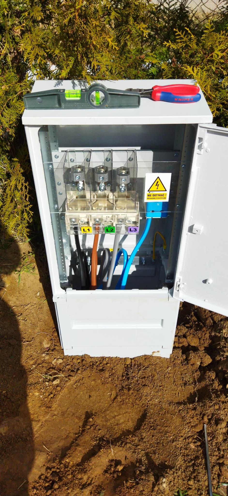 Otwarta skrzynka elektryczna z podłączonymi przewodami oznaczonymi L1, L2, L3, poziomica i szczypce leżą na górze, widoczne ostrzeżenie o napięciu.