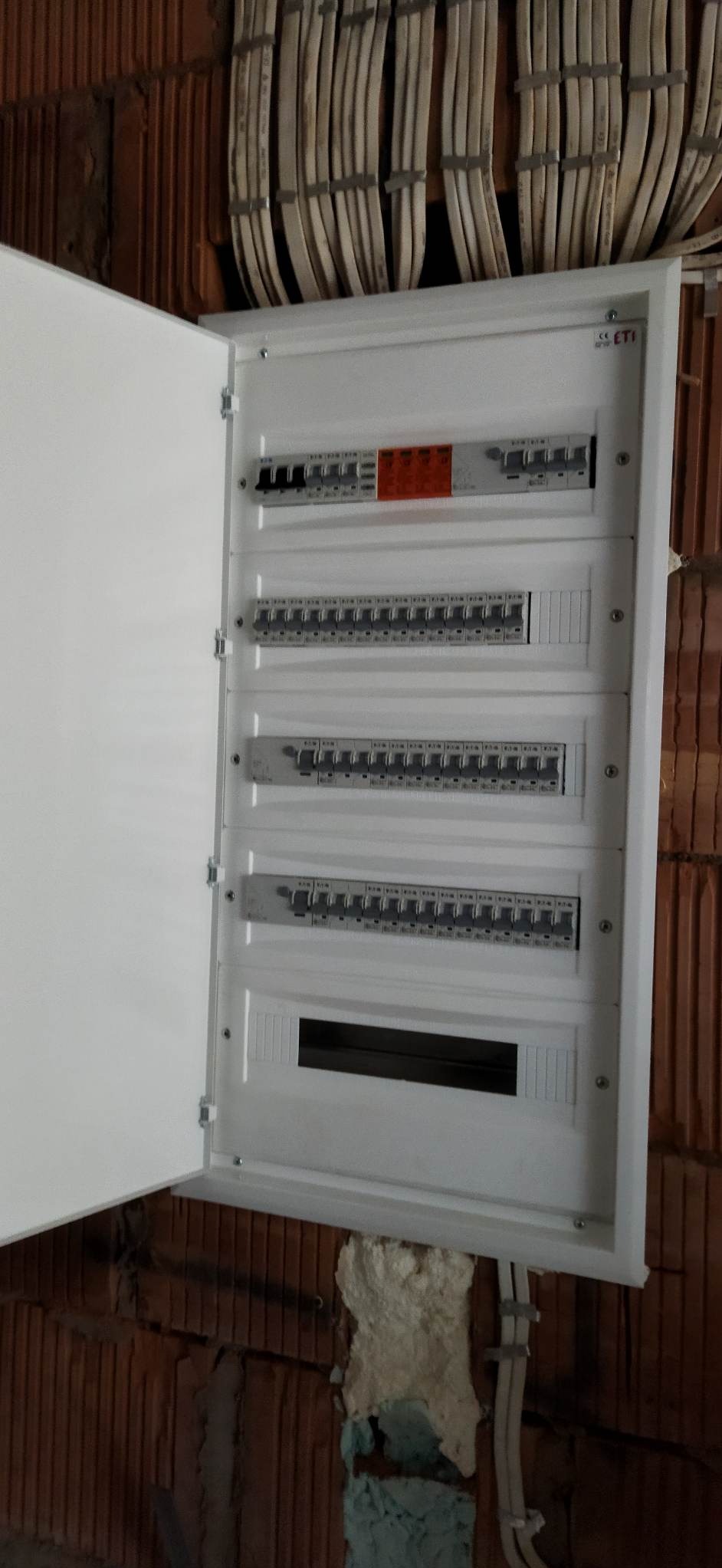 Otwarta skrzynka z bezpiecznikami zamontowana na ceglanej ścianie z widocznymi przewodami elektrycznymi. Widoczne zabezpieczenia różnicowoprądowe i nadprądowe.