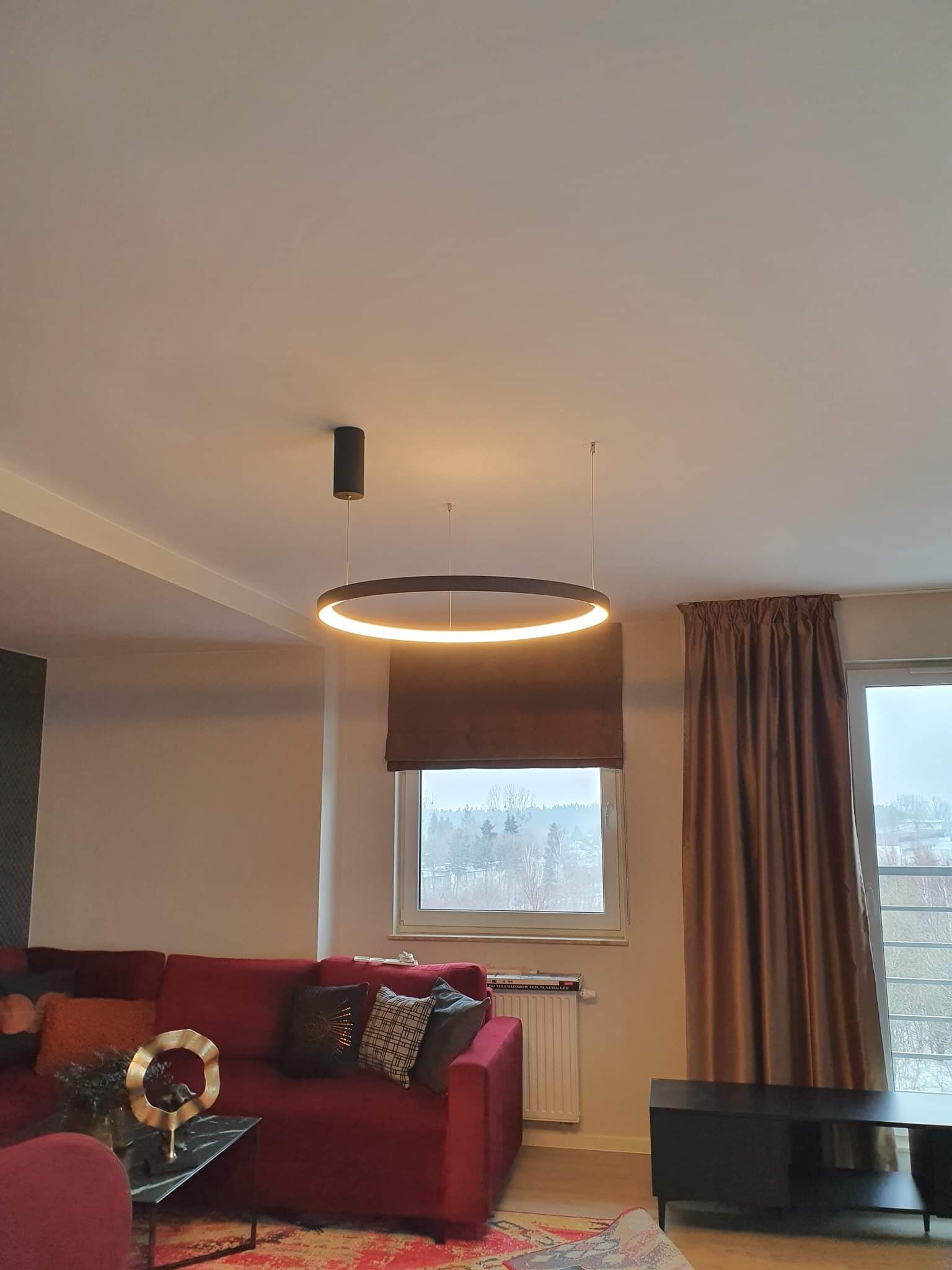Nowoczesny okrągły żyrandol LED zawieszony w salonie nad czerwonym narożnikiem, widok na okno z brązowymi zasłonami i roletami rzymskimi.