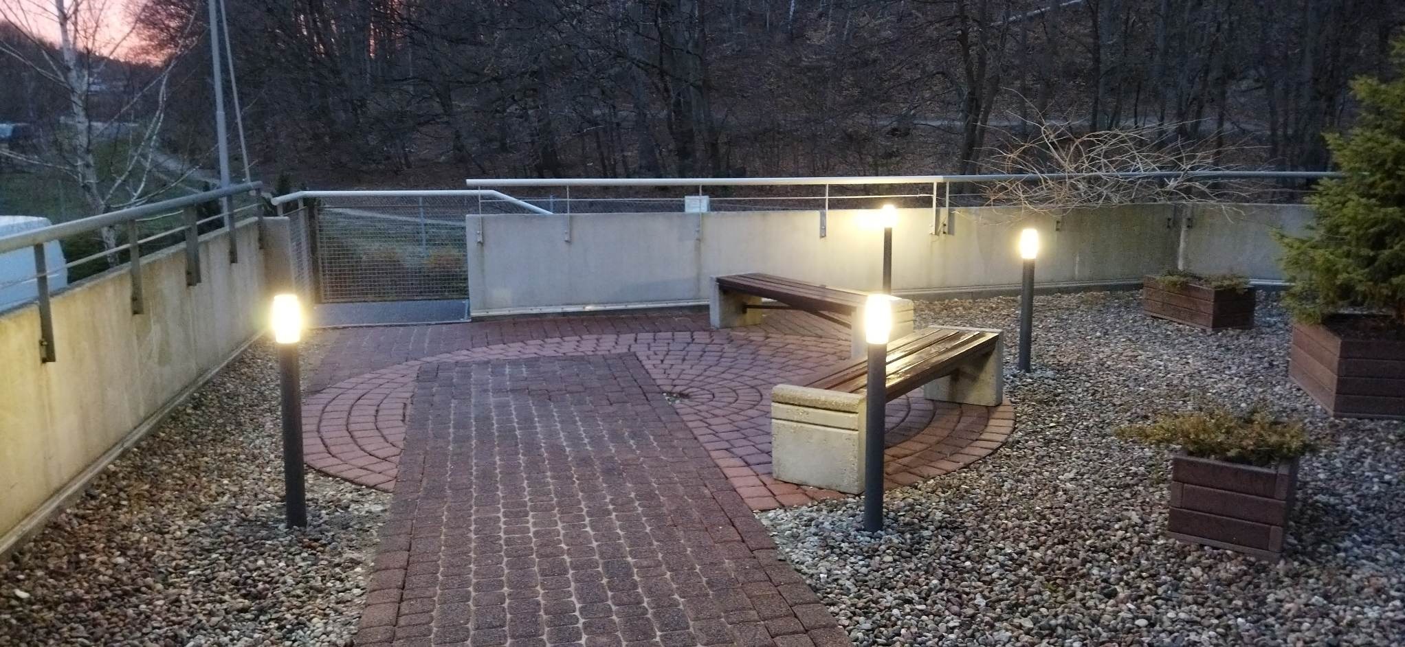 Oświetlony plac z brukowaną ścieżką, ławkami z betonu i drewna oraz lampami stojącymi, otoczony żwirem i zielenią w donicach.