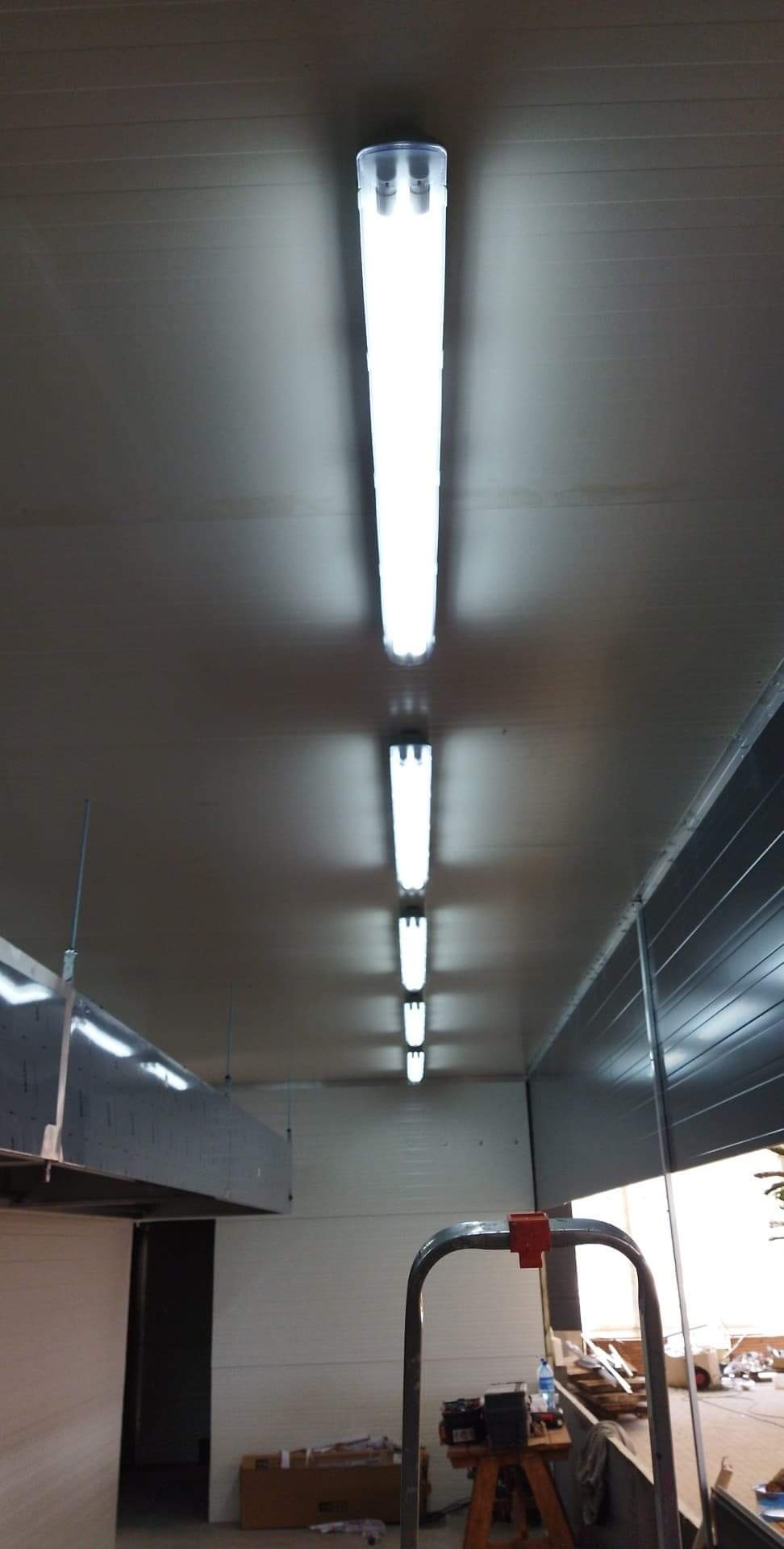 Równy rząd lamp LED zamontowanych na suficie w hali produkcyjnej, widoczna drabina i elementy wentylacji.