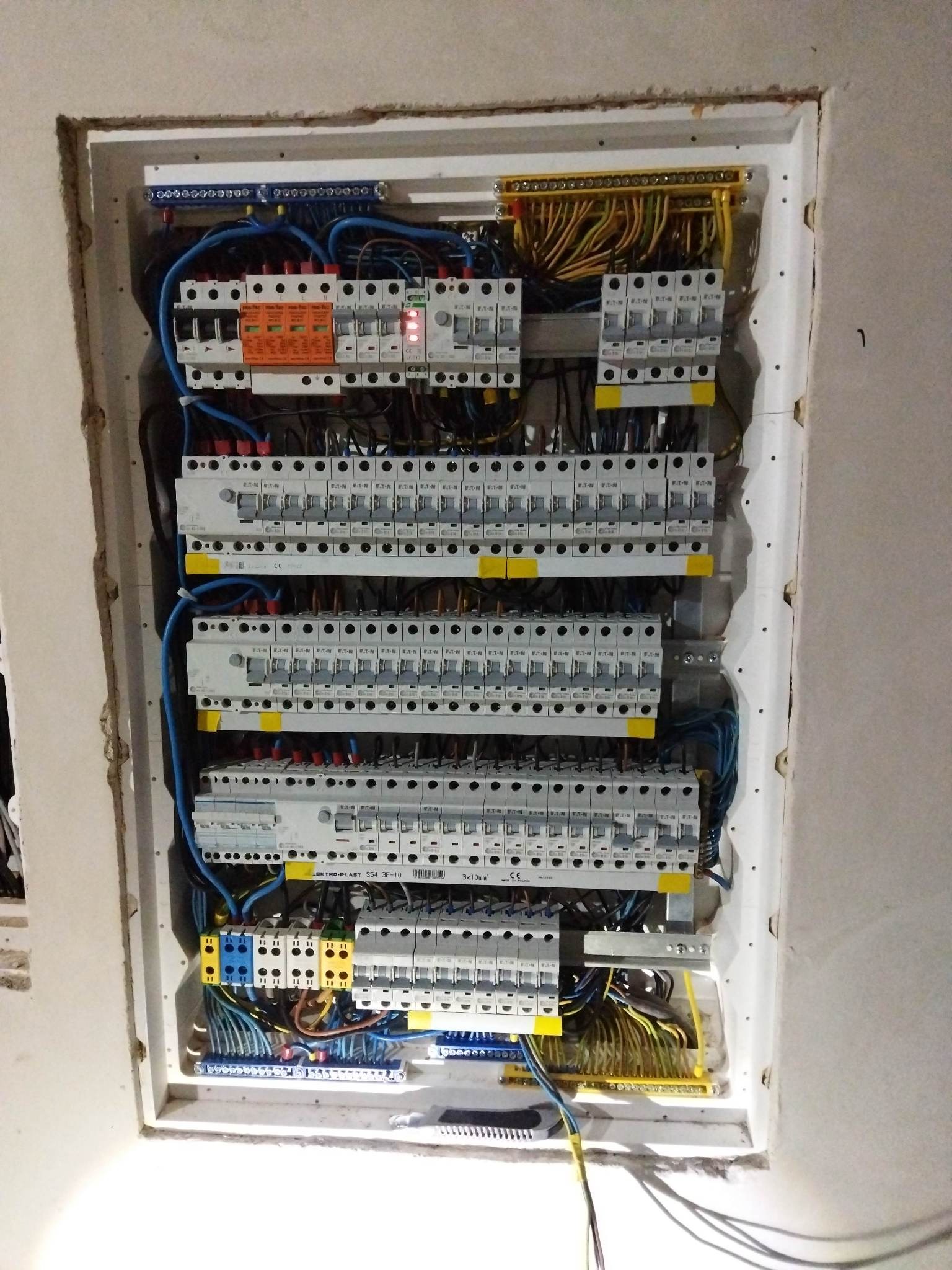 Rozdzielnica elektryczna wnękowa z licznymi bezpiecznikami, zabezpieczeniami różnicowoprądowymi i przewodami, widoczne oznaczenia na urządzeniach.