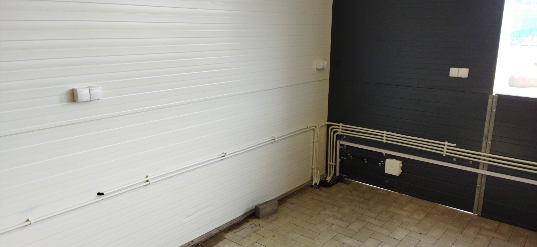 Białe i czarne panele ścienne z widocznymi rurami instalacji elektrycznej przy podłodze i puszkami elektrycznymi na ścianach, układane wzdłuż krawędzi pomieszczenia.