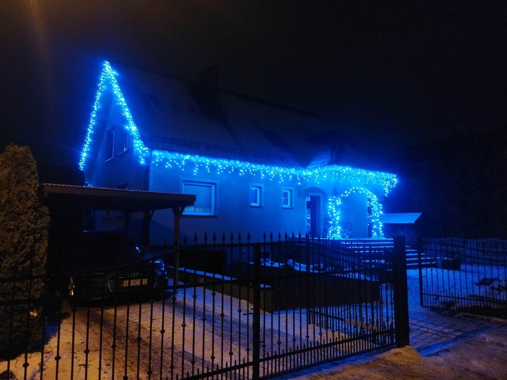 Dom jednorodzinny udekorowany niebieskimi lampkami świątecznymi, widok nocny zza metalowego ogrodzenia.